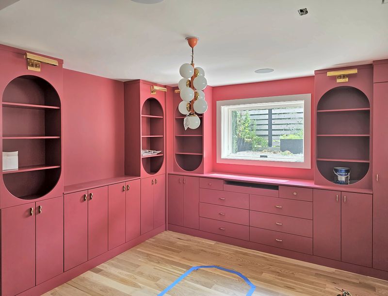 Raspberry-red painted library wall with arched open shelving niches, brass picture lights, lower cabinets and a bank of drawers under a window
