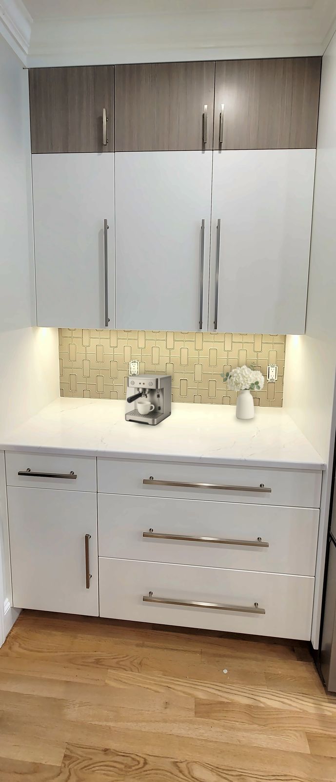 Two-tone coffee bar with white slab drawers and doors below, wood-grain upper cabinets above and under-cabinet lighting on a tile backsplash, styled with an espresso machine and a vase of flowers