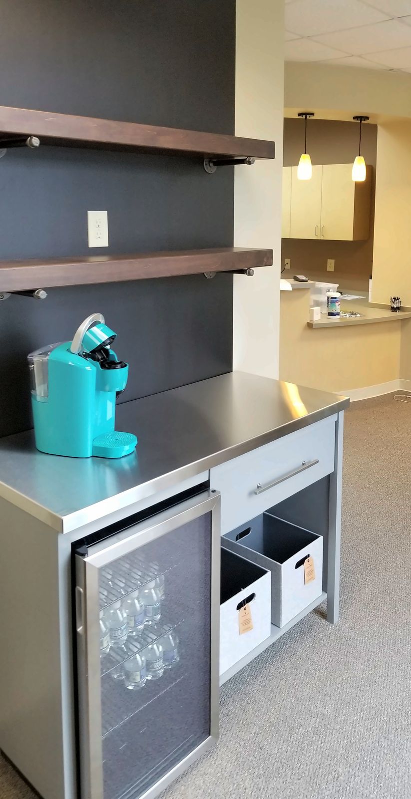 Coffee bar with a stainless-steel top over a wine fridge and open storage, with two walnut floating shelves on a dark wall
