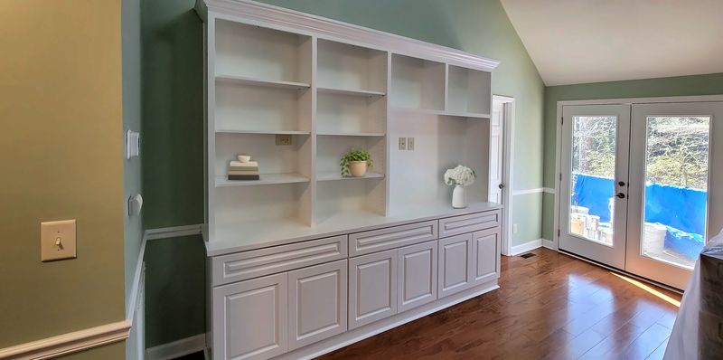 White built-in bookcase wall with open adjustable shelves above raised-panel lower cabinets, styled with books, a plant and a vase of flowers
