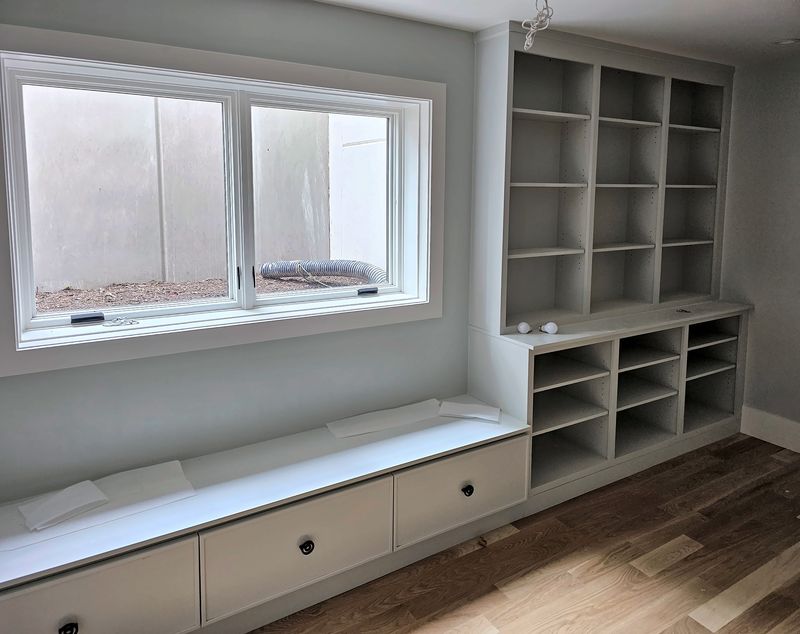 White window seat with drawers beneath a wide window, next to a built-in open bookcase