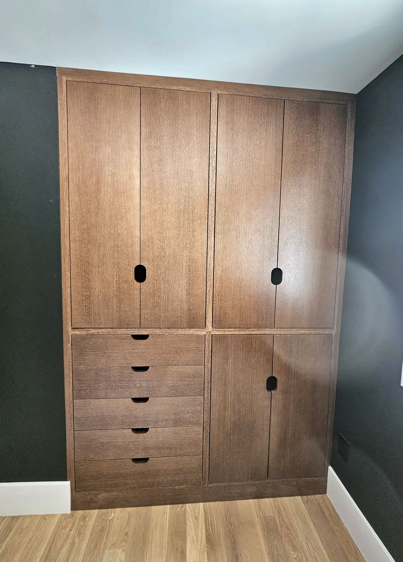 Floor-to-ceiling stained oak wardrobe with flush doors, round finger-pull cutouts and a bank of drawers, against a dark wall