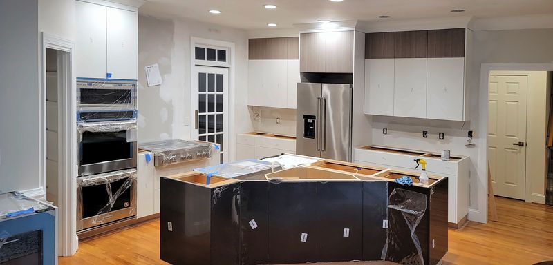 Kitchen during installation with white perimeter cabinets, stacked wall ovens and a dark island waiting for its top
