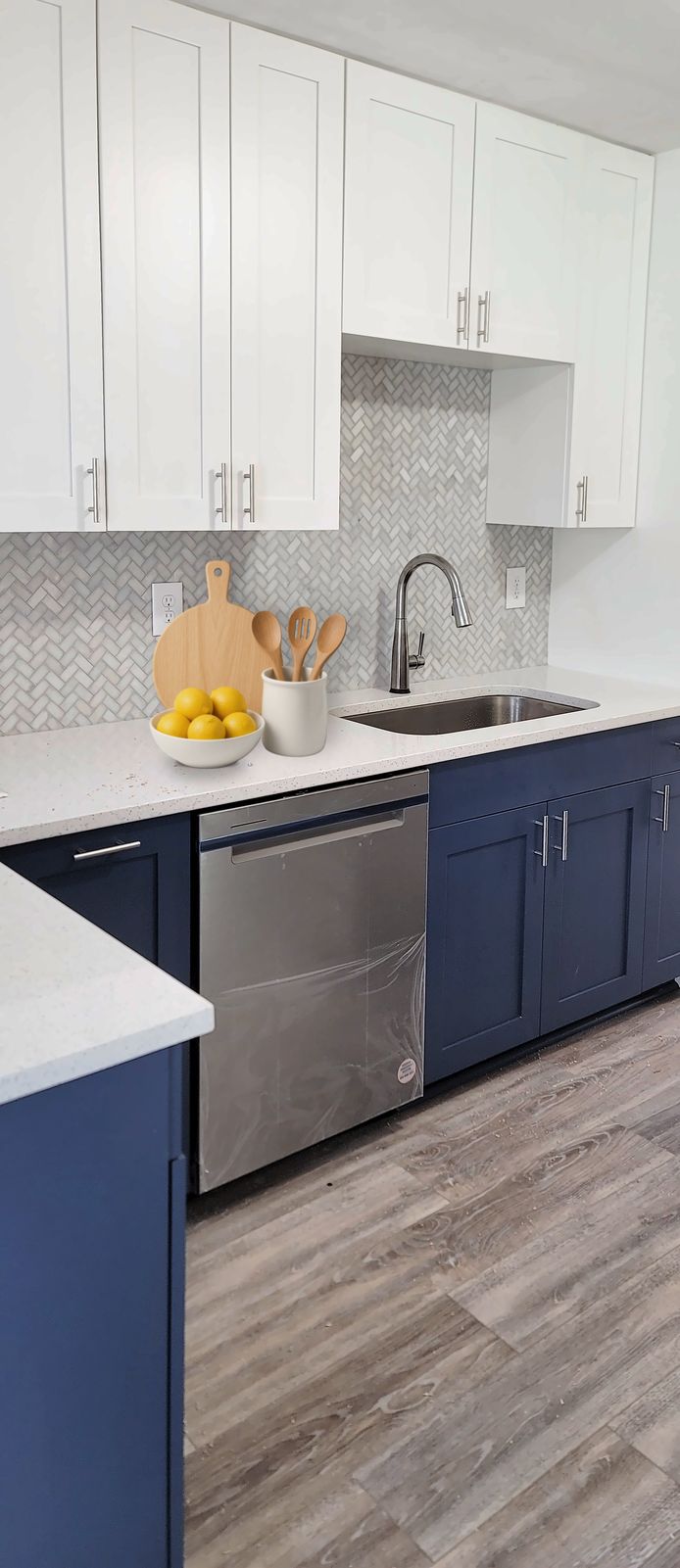 Galley kitchen with navy shaker lower cabinets, white shaker upper cabinets to the ceiling, a white counter and a stainless dishwasher, with a cutting board, utensils and lemons by the sink