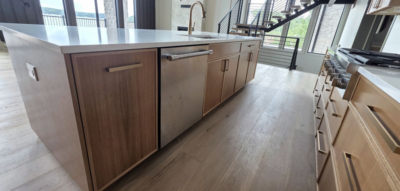 Long light oak kitchen island with flat-panel drawers and doors, a paneled dishwasher opening and a white quartz top, with oak base cabinets opposite, in Macon, NC