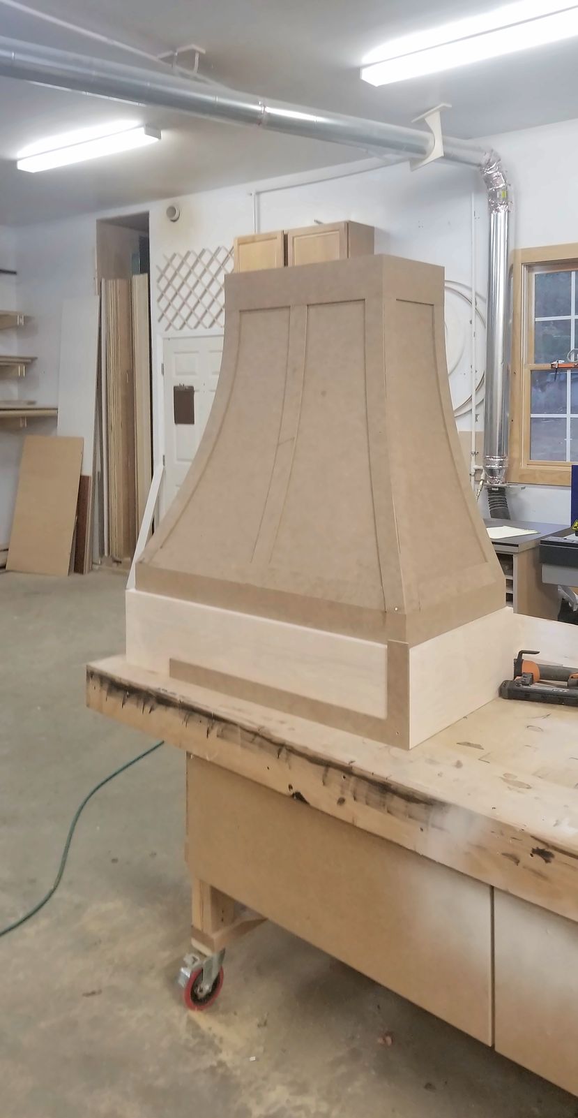 Unfinished wood range hood with flared sides standing on a workbench in the shop
