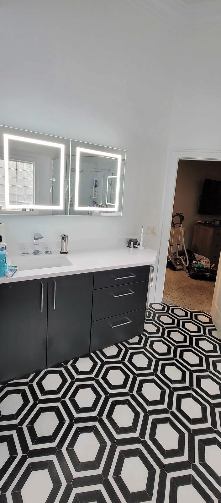 Black flat-slab vanity with a white top and two lighted mirrors, above a black and white hexagon tile floor