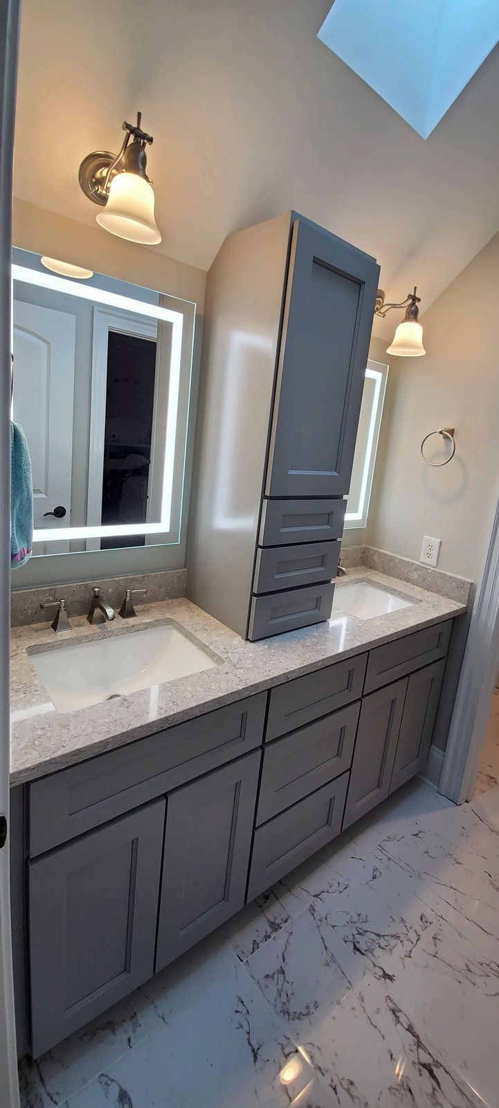 Gray shaker double vanity with a tall center linen cabinet between two lighted mirrors, over a marble-look floor