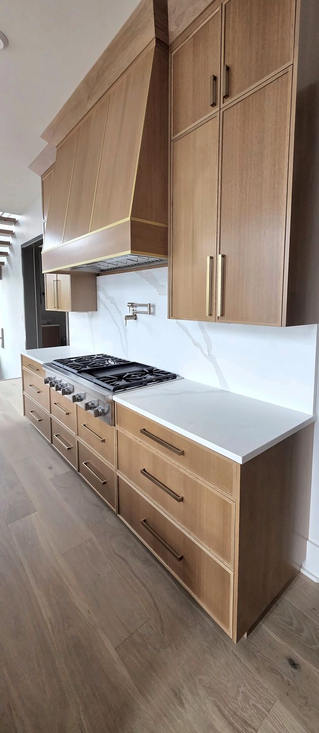 Light oak range wall in a Macon, NC kitchen: tall oak upper cabinets with brass pulls, an oak range hood with brass trim, and oak drawers under a white quartz counter