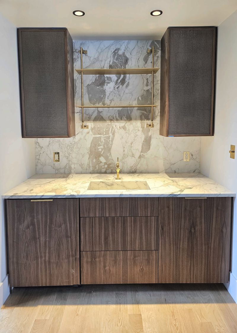 Walnut cabinetry built into a niche, with slab drawer fronts, a white marble counter and full-height marble backsplash, a brass wall shelf and two walnut-framed upper cabinets with woven mesh doors