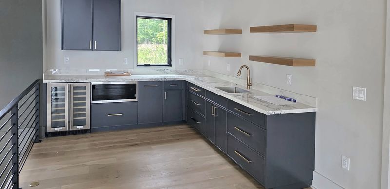 Navy kitchenette in a Macon, NC loft with navy flat-panel cabinets, brass pulls, a white marble-look counter, a wine fridge and light oak floating shelves